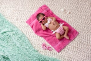 A baby, wearing sunglasses and a crochet bikini, lies on a pink towel on a textured surface. Nearby are striped flip-flops and small beach trinkets. A light green fabric drapes across the bottom left corner, adding a perfect summer touch to remember those future summer girl names or boy names for next season.