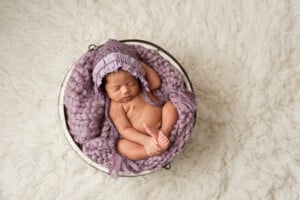 A newborn baby wearing a purple knit bonnet is sleeping in a curly position inside a round white basket filled with a purple knit blanket. The basket is placed on a cream-colored, fluffy rug. Looking serene and precious, she's sure to inspire parents seeking unique girl names that start with X.