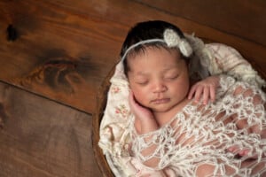A sleeping baby with short black hair rests in a round wooden bowl, wrapped in a white knit blanket. The baby has a delicate headband with a small flower decoration and is lying on floral patterned fabric. The background is a wooden surface, evoking the charm of biblical girl names like Ruth or Esther.