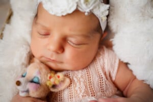 An infant girl, possibly with a one-syllable name like Grace or Jane, is wearing a white headband with flowers and a lace outfit. She is peacefully sleeping while holding a small, plush toy of a deer. The baby is nestled in a soft, fluffy white blanket.