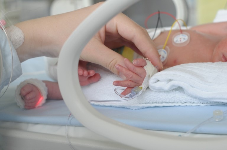 An infant lies in an incubator with medical wires attached to its chest and left hand. An adult hand, likely that of a NICU mom, gently holds the baby's right hand. The infant is covered in a white cloth, and a tube is connected to the baby's hand with medical tape and bandage.