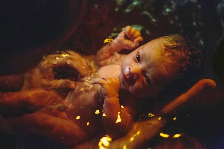 A newborn baby is held in dimly-lit water, with golden reflections on the surface. The baby has wet hair and looks up with eyes partially open, evoking a serene moment synonymous with the gentle experience of an orgasmic birth. The infant appears to be in the hands of an adult, partially submerged.