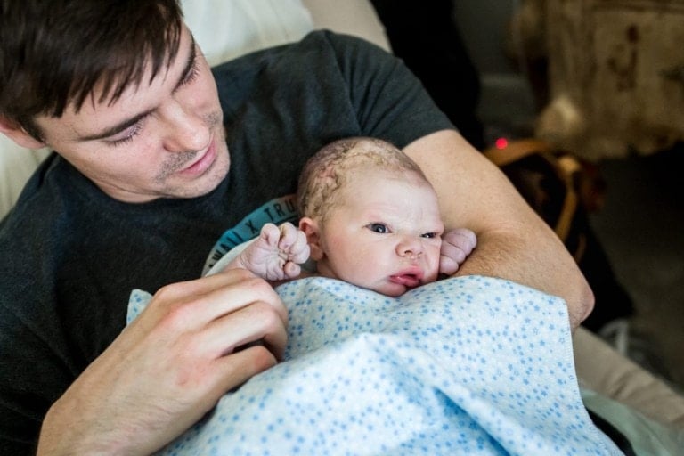 A man with short brown hair sits while holding a newborn baby wrapped in a light blue blanket with blue star patterns. The baby is resting against the man's arm, looking up with wide eyes, and he exudes the joy of becoming a dad.