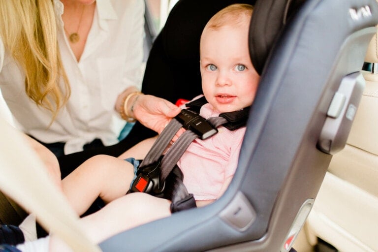 A baby with light hair sits in a Nuna baby car seat, looking at the camera. An adult, partially visible and wearing a white shirt, adjusts the car seat straps to secure the baby. The car interior is visible in the background.