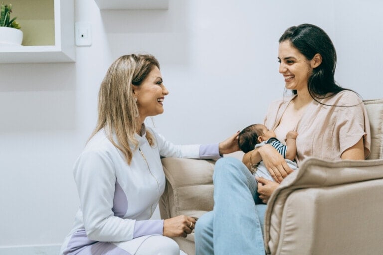 A woman is seated on a beige chair, breastfeeding a baby while smiling. Another woman, wearing a medical uniform, kneels beside her and smiles, placing a hand on her arm. They appear to be in a clinical or office setting with white walls. This scene raises the question: what is an IBCLC?