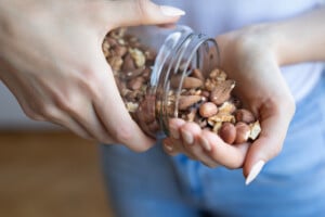 A person is pouring a mixture of nuts, including almonds, hazelnuts, and walnuts, from a small glass jar into their cupped hand. The background is blurred, and the person is wearing a light-colored top and blue pants—perfect postpartum snacks for new moms looking for a nutritious boost.