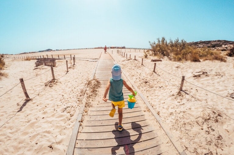 A child walking on a wooden boardwalk toward the beach, holding a blue bucket and shovel. Emphasizing beach safety, the child is wearing a light blue shirt, yellow shorts, and a white hat. The sky is clear and the surrounding area is sandy with some sparse vegetation.