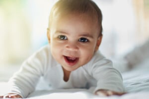 A baby named Rowan lies on its stomach on a white surface, facing the camera with a smile. The baby is wearing a white outfit. The background is softly blurred with natural light, creating a warm and gentle atmosphere.