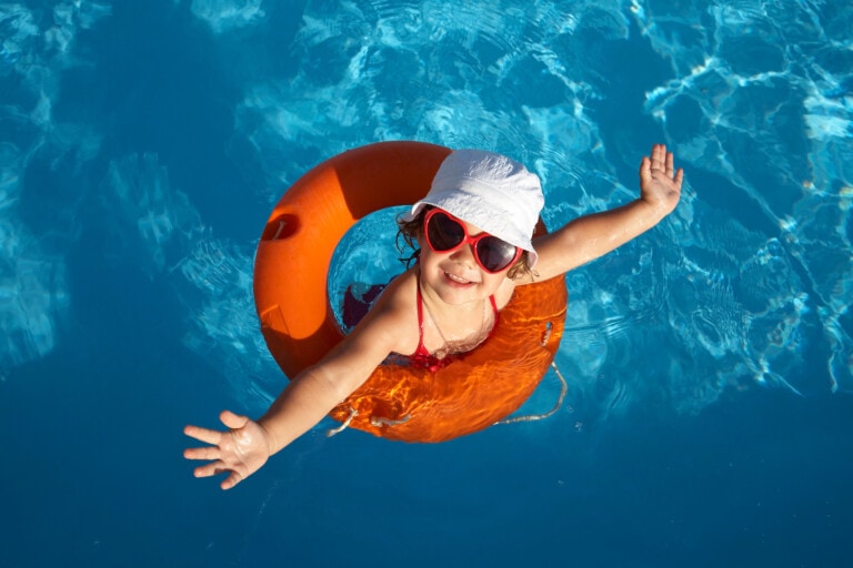 A child is floating in a swimming pool using an orange inflatable ring. The child is wearing a white hat, red sunglasses, and a red swimsuit, with arms outstretched while smiling up at the camera. This snapshot perfectly captures one of the fun summer activities for kids in clear, blue water.