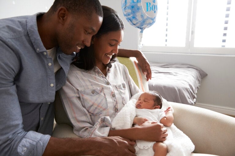 A man and a woman sit closely together with a newborn baby in their arms. The woman is holding the baby, just brought home from the hospital, and the man has his arm around them both. A blue balloon with the words "It's a Boy!" is seen in the background. They appear to be in a bedroom.