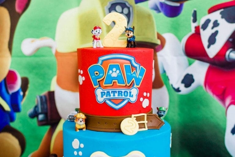 A two-tiered cake for a Paw Patrol Birthday Party is decorated with small figurines of characters from the show. The top layer is red with the "Paw Patrol" logo and a number 2, while the bottom layer is blue and features a brown belt decoration around it.