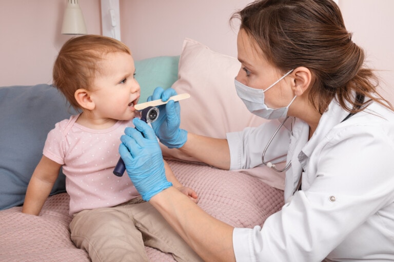 A healthcare professional wearing a mask and gloves uses a tongue depressor and otoscope to examine the red throat of a baby sitting on a bed. The baby, dressed in a pink shirt, appears cooperative during the examination. Various pillows are in the background.