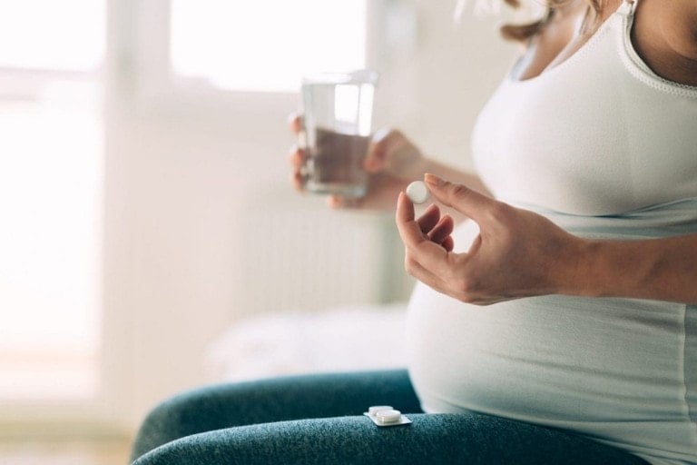 A pregnant person sits on a bed holding a glass of water in one hand and a tablet in the other, seeking pregnancy heartburn relief. Wearing a white tank top and blue leggings, they hope to find comfort from heartburn during pregnancy. Additional tablets rest on their leggings near the bottom of the image.