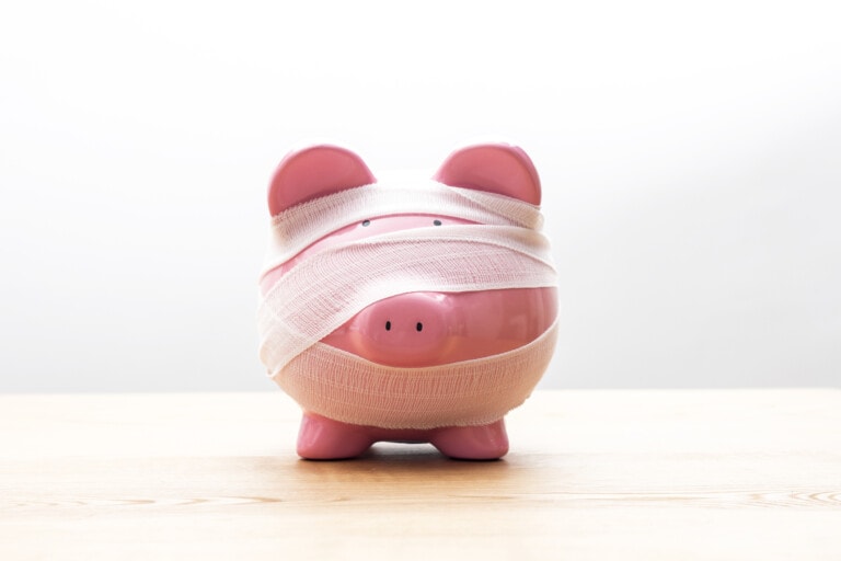 A pink piggy bank, wrapped in white bandages like an emergency fund on the mend, covers most of its body and face, leaving only the snout visible. The bandaged piggy bank sits on a light wooden surface against a white background.