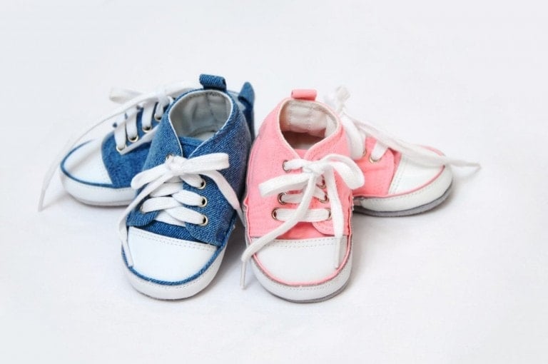 Four baby shoes are displayed against a white background. Two shoes are blue with white laces, and two are pink with white laces, subtly hinting at gender swaying. The shoes feature a casual sneaker style designed similarly for both colors.