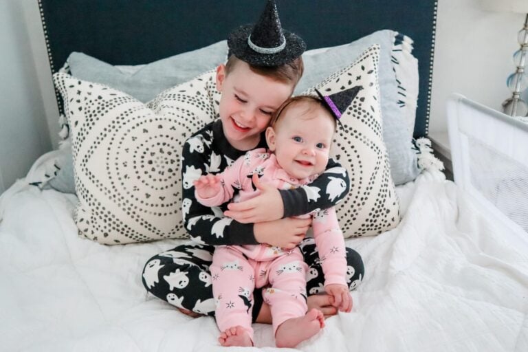 A young child and a baby sit on a bed dressed in charming halloween pajamas. The older child, wearing black and white ghost-patterned pajamas and a witch hat, hugs the baby, who is snug in pink pajamas with a small hat. They are cozily positioned in front of two patterned pillows.