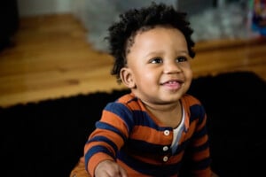 A young boy with curly hair and wearing a striped orange and blue shirt is sitting on a black rug. The child, potentially named Victor or Vincent from popular V names for boys, is smiling and looking slightly upwards. The background includes a wooden floor and some indistinct objects.
