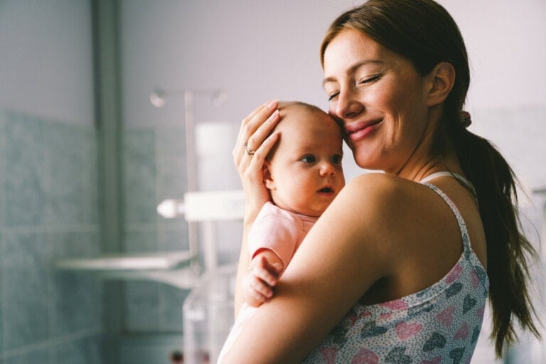 A woman holds a baby close to her chest, smiling contentedly. The baby is looking off to the side, while the woman seems happy and serene—exemplifying Dr. Sears' attachment parenting principles. They are in a room with soft natural light and light-colored walls. The woman is wearing a tank top with a heart pattern.