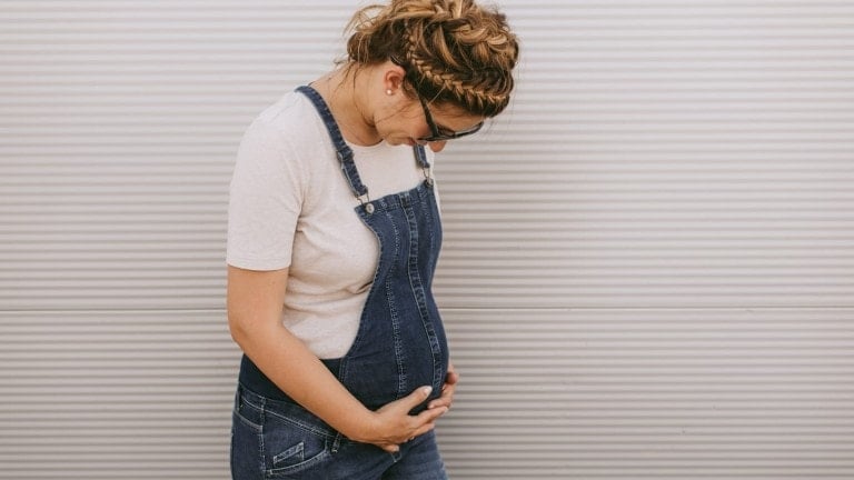 A pregnant woman stands sideways against a white, textured background. She is looking down and has her hands gently placed on her baby bump. She is wearing a white T-shirt and denim overalls, with her hair styled in a braid, possibly listening to the Say to a Pregnant Person podcast.