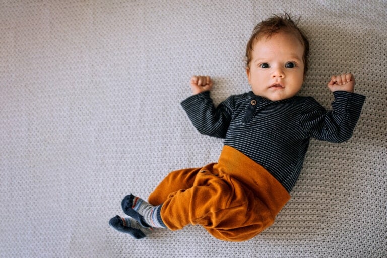 A baby wearing a striped long-sleeve shirt, orange pants, and striped socks lies on a textured white blanket. The baby, who looks like they could have one of those classic German names, has brown hair and wide eyes, looking up at the camera with both arms bent at the elbows.