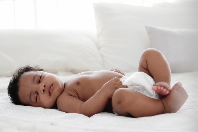 A baby is lying on their back asleep, wearing only a diaper. The baby is partially covered with a white blanket, and the background features white pillows and bedding. The scene is softly lit, creating a calm and peaceful atmosphere—ideal for those seeking baby sleep tips.
