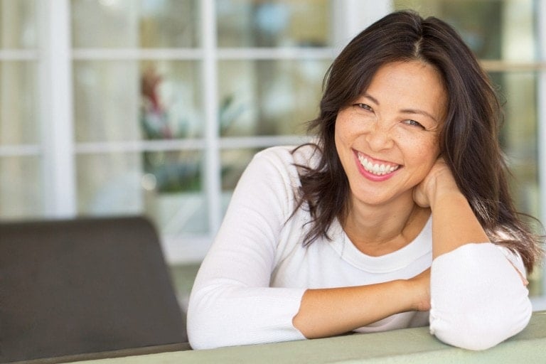 Smiling woman with medium-length dark hair, wearing a white long-sleeve top, leaning on a surface with her right elbow and resting her head on her right hand. The serene mood is perfectly captured against the background of large white-framed glass windows.