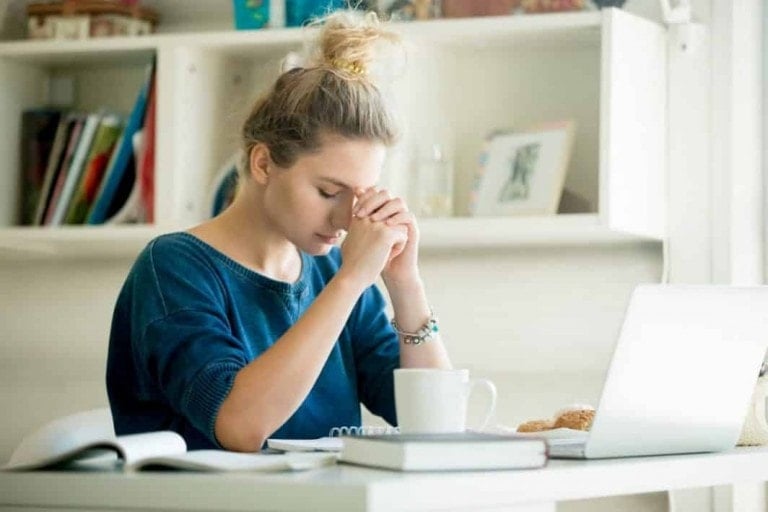 A woman sits at a desk with her eyes closed and hands clasped near her face, appearing stressed. She is in front of an open laptop, with books, a coffee mug, and a pastry nearby. Shelves with books and decorations are visible in the background as she tries to find ways to relieve stress.