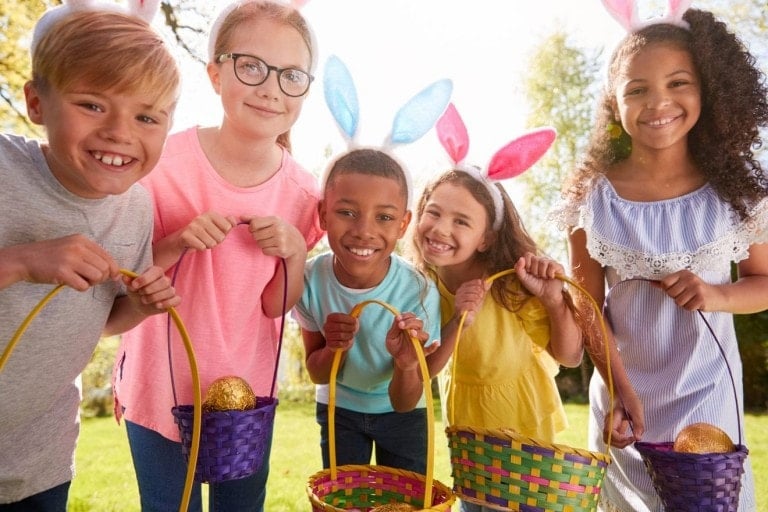 Five children are outdoors, smiling and holding colorful Easter baskets. They are wearing headbands with bunny ears, likely inspired by some creative Easter basket ideas. The group appears to be engaged in an Easter egg hunt. The background shows trees and grass under a bright sunny sky.