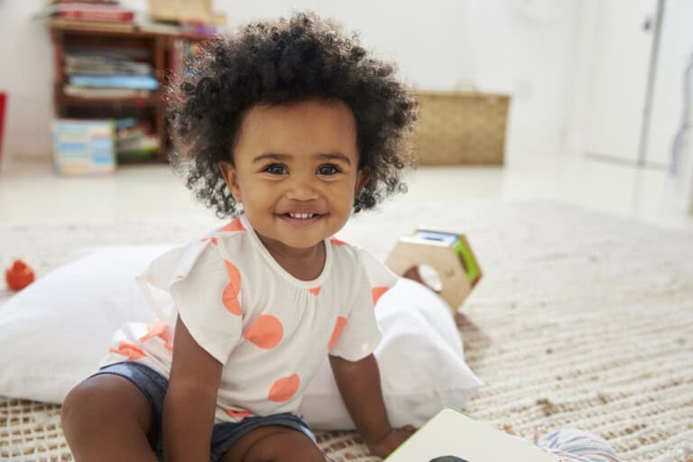 A young girl with curly hair and a big smile sitting on a rug, reflecting the joy of reaching new 16-month-old milestones. The child is wearing a white shirt with orange polka dots and denim shorts. There are toys, a pillow, shelves of books, and a basket in the background, perfect for activities for 16-month-olds.
