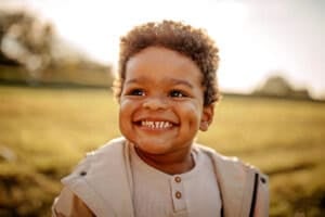 A 22-month-old child with curly hair is smiling broadly, showing dimples, while standing outdoors. The toddler is wearing a light-colored jacket and shirt. The background is a blurred natural setting with sunlight illuminating the scene, perfectly capturing a moment of joyful discovery.