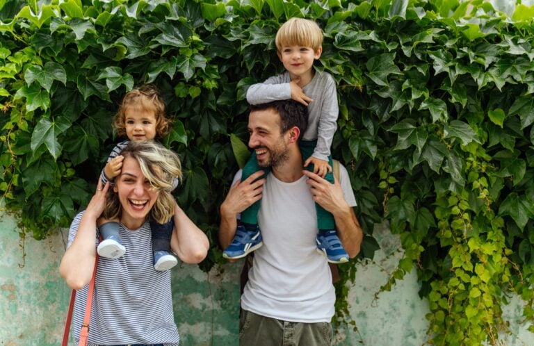 A woman and a man are standing in front of a lush green wall, each carrying a child on their shoulders. The woman is smiling widely with a toddler on her shoulders, and the man is laughing, holding a young child on his shoulders. Their joyful expressions reflect the warmth found in Baumrind's parenting styles.