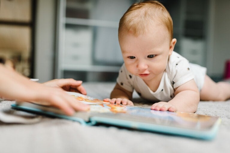 A baby lies on their stomach on a bed, intently looking at one of the best baby books. An adult's hands are visible holding the book open. The baby is wearing a white onesie with small black designs. The background is out of focus, suggesting a bedroom setting.