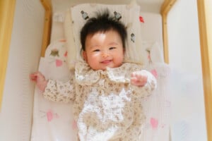 A baby girl named Kayla wearing a light-colored, patterned outfit lies on a soft surface. The baby is smiling and raising one arm. The surroundings include fabric with bird and animal-themed prints. The bright light suggests a warm and cheerful atmosphere, perfect for capturing precious moments.