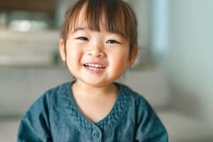 A young child with straight brown hair, bangs, and a blue shirt smiles while sitting indoors. The softly blurred background highlights a neutral-colored couch and wall—perfect for capturing special 2-year-old milestones.