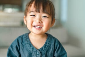 A young child with straight brown hair and bangs is smiling at the camera. The child, possibly named after u girl names like Uma or Ursula, is wearing a blue-buttoned shirt. The background is slightly blurred and appears to be an indoor setting.