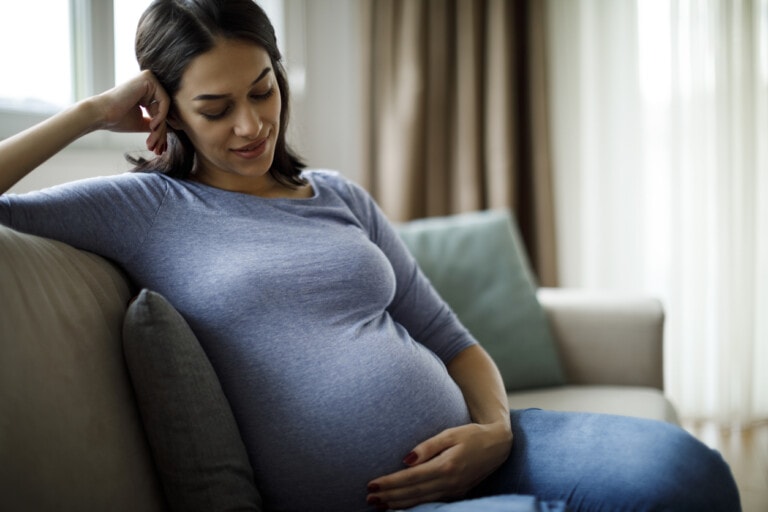 A pregnant woman with dark hair is sitting on a beige couch, resting her head on her hand. She is wearing a blue long-sleeve top and jeans, appearing to look down and smile slightly. In the background, there are a window and curtains. Her expression reflects the peaceful contemplation of pregnant mom thoughts.