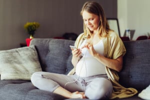 A pregnant woman sits cross-legged on a gray couch, smiling as she looks at her smartphone. She is wearing a white top, light gray leggings, and earphones, likely enjoying her birth playlist. In the background, there is a blurred view of a table with a yellow flower vase and other household items.