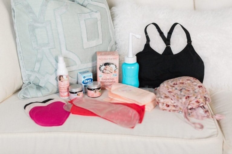 A collection of postpartum must-haves arranged on a soft beige couch includes a black nursing bra, Perineal Bath Herbs, a Perineal Care Spray, Earth Mama Bottom Balm, QuickClean Micro-Steam Bags, maternity pads, cloth wipes, a peri bottle, and a floral bag.