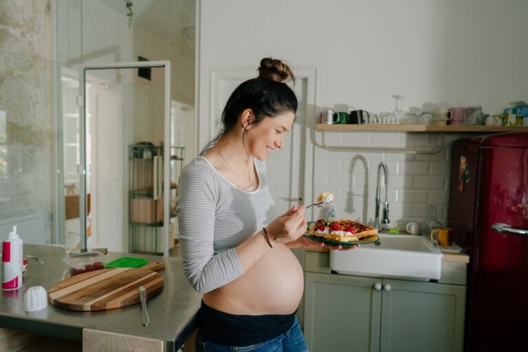A pregnant woman with her hair in a bun is standing in a kitchen, eating for two from a plate of waffles topped with fruit and whipped cream. She is wearing a grey striped top and jeans. The kitchen has a red refrigerator, white sink, and various kitchen items.