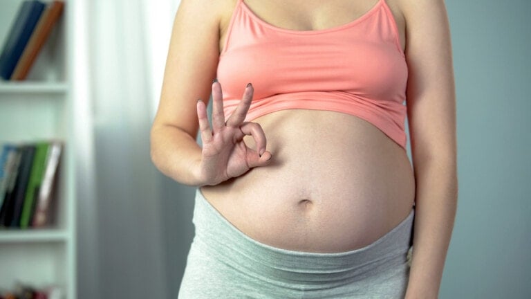 A pregnant person stands indoors with a hand resting on their belly, revealing the truths about the third trimester. They are wearing a coral-colored sports bra and light gray leggings. Their left hand makes an okay gesture, while the background features shelves with books and a bright window.
