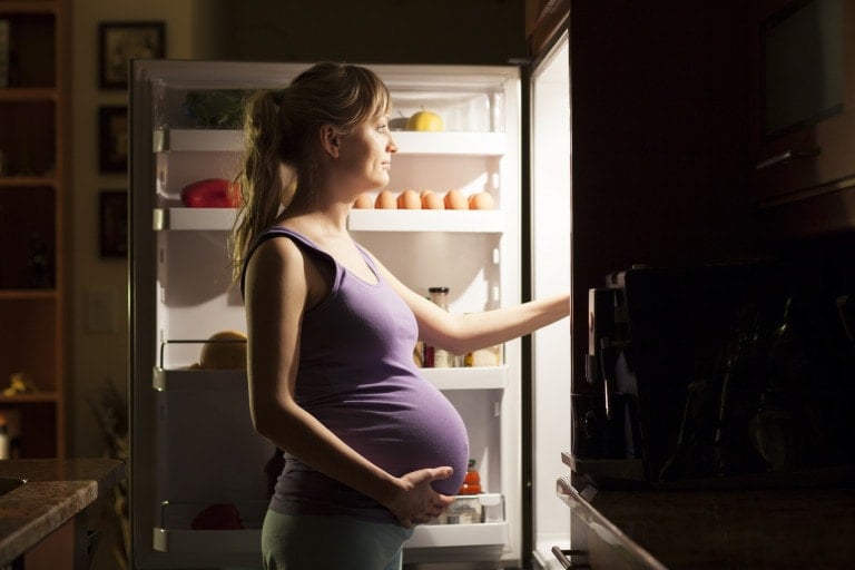 A pregnant woman stands in front of an open refrigerator, looking inside. She is holding her belly with one hand while reaching into the fridge with the other, contemplating what to eat during pregnancy. The kitchen is dimly lit, and the light from the refrigerator illuminates her face.