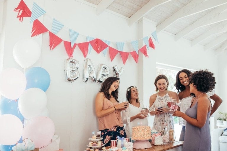 Five women are gathered at the baby shower, standing around a wooden table adorned with a cake and various snacks. The room is decorated with balloons, and a banner spelling "BABY" hangs on the wall behind them, setting the perfect scene for celebrating baby showers.