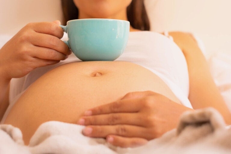 A pregnant woman is lying down, holding a light blue cup near her belly—likely one of her favorite drink swaps. Her other hand is resting on her stomach, and she is covered with a white blanket. She is wearing a white tank top.