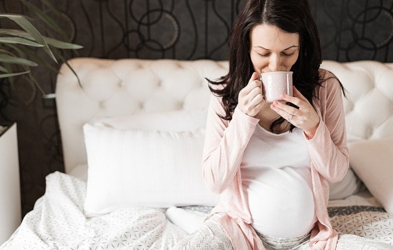 A pregnant woman with long dark hair sits on a bed, wearing a light pink cardigan and white top. She holds a pink mug with both hands, seemingly drinking red raspberry leaf tea. The bed has white and patterned grey bedding, and there is a plant in the background, adding to the serene atmosphere of her pregnancy journey.