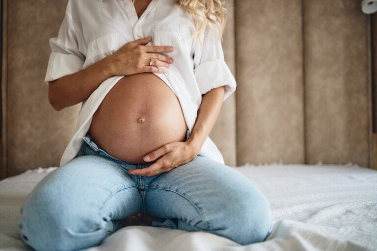 A pregnant person in a white shirt and blue jeans is sitting on a bed, holding their bare belly with both hands—a perfect moment highlighting second-trimester must-haves. The background includes a beige padded headboard.
