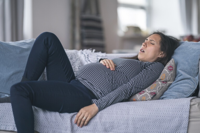A pregnant woman in a striped shirt and dark pants is reclining on a couch with her head resting on a pillow. She appears to be in discomfort, with her hand on her stomach and her face showing signs of strain. Pregnancy can be a big deal when you're constantly dealing with such discomforts. The background includes some furniture and home decor items.