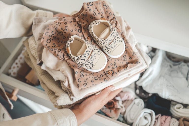 A pair of toddler shoes with a leopard pattern is placed on top of a stack of folded clothes. The clothes and shoes are being held by a person who's in the midst of packing baby clothes near an open drawer filled with neatly organized garments.
