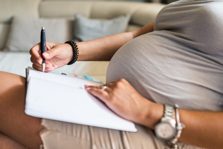 A pregnant woman is sitting on a couch and writing in a notebook, creating a birth plan. She is holding a pen in her right hand and resting the notebook on her lap. She is wearing a gray shirt, beige shorts, a bracelet, and a watch. The setting appears to be a home living room.