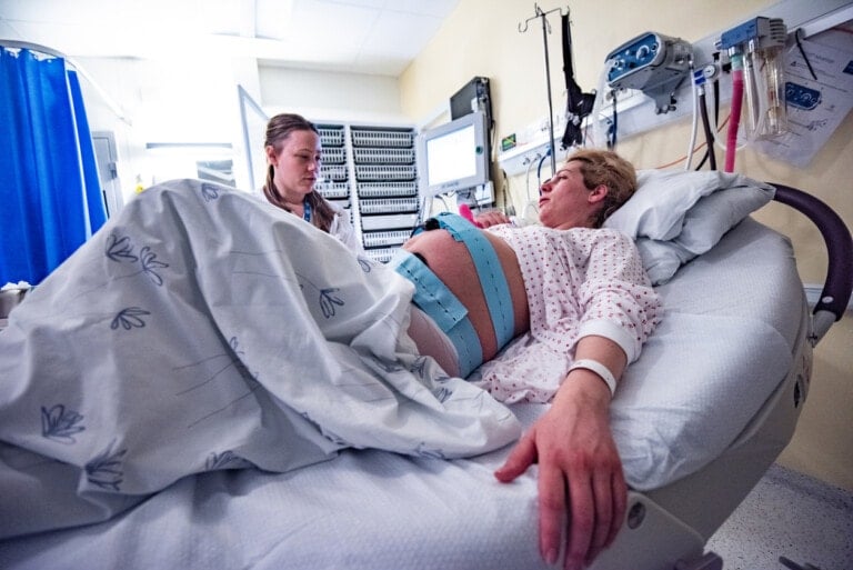 A pregnant woman lies in a hospital bed with a fetal monitor belt around her abdomen, under observation for potential umbilical cord prolapse. A healthcare worker is seated beside her, carefully monitoring her condition. Medical equipment and monitors are visible in the background.