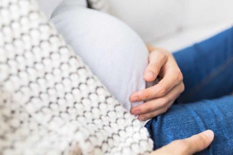 A close-up of a pregnant person cradling their belly. The individual is wearing a white, ribbed shirt, blue jeans, and a gray knitted cardigan. Their hands gently rest on the stomach, as if shielding against common pregnancy myths, emphasizing the beauty of this natural journey.